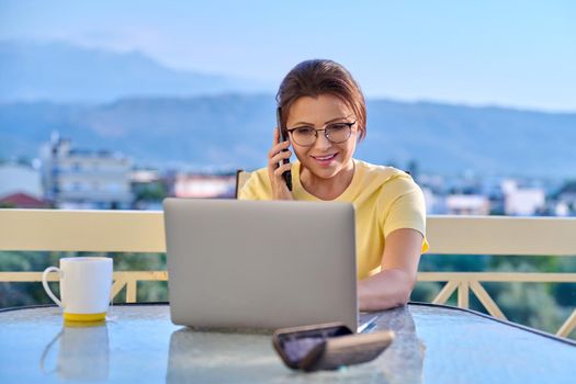 Middle aged woman working at home on terrace with laptop smartphone. Business female talking with client, colleague on phone, looking at laptop screen. Freelance, remote work, technology in business