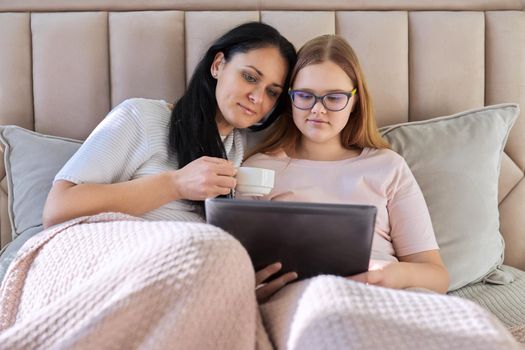 Mother and teenage daughter resting at home together, use digital tablet. Woman and teen girl lying in bed, looking at screen, movie video. Family, parent teenager relationship, vacation, technology