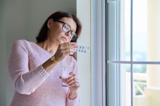 Middle-aged woman with pills and glass of water in her hands, at home near window, copy space. Mature women's health, pharmacology, vitamins, supplements, therapy, treatment