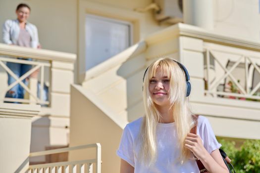 Portrait of a girl student of a teenager 17, 18 years old in headphones with a backpack going to college, to high school. Porch of the house, mom missing daughter background.