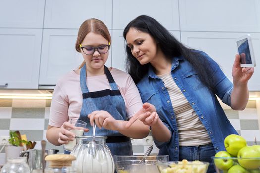 Mom and teen daughter preparing apple pie together, at home in kitchen. Woman with digital tablet, recipe, video call, technology in daily life. Family, parent teenager relationship, lifestyle