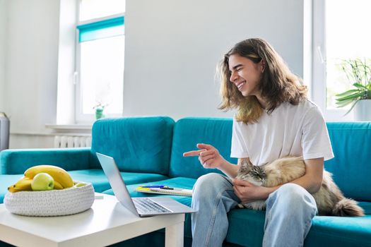 Guy teenager studies at home online with laptop and pet cat. Male 16, 17 years old sitting on couch looking at webcam, talking, using video call. E-learning, virtual lessons, technologies in education