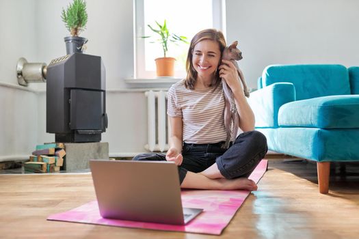 Middle-aged smiling woman with laptop and cat on the floor. Positive female sitting on a yoga mat in a lotus position with a pet in her arms looking at a laptop screen using a video call