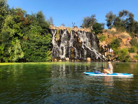 Monastyrsky Island, Dnipro, Ukraine. A group of tourists descends to artificial waterfall. A girl on sup rafting down the river. The waterfall is surrounded by trees against a blue sky without clouds