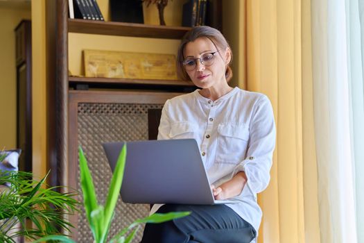 Middle-aged woman psychologist in an office with laptop. Portrait of professional female counselor, psychiatrist, therapist, teacher sitting on chair near window. Technology, medicine, people