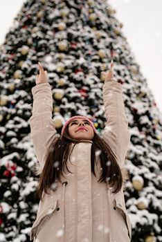 Merry Christmas and happy New Year. Happy Woman in warm winter clothes standing by the big christmas tree outdoors, arms up, snow falling