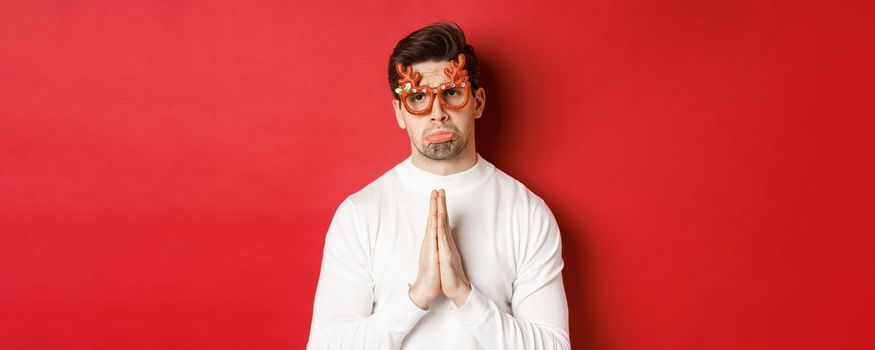 Image of handsome man in christmas party glasses, begging for help or apologizing, need a favour, standing over red background.