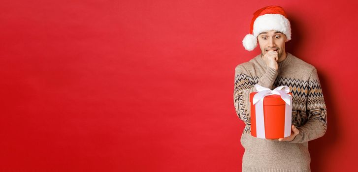 Image of worried guy in santa hat and sweater, looking indecisive at christmas gift, standing against red background.