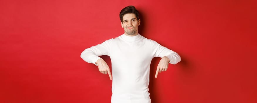 Portrait of attractive, skeptical young man in white sweater, grimacing and pointing fingers down, disagree and dislike something, standing over red background.