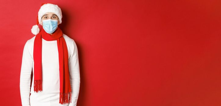 Concept of new year, coronavirus and holidays. Cheerful man celebrating christmas and social distancing, wearing medical mask, santa hat and scarf, standing over red background.