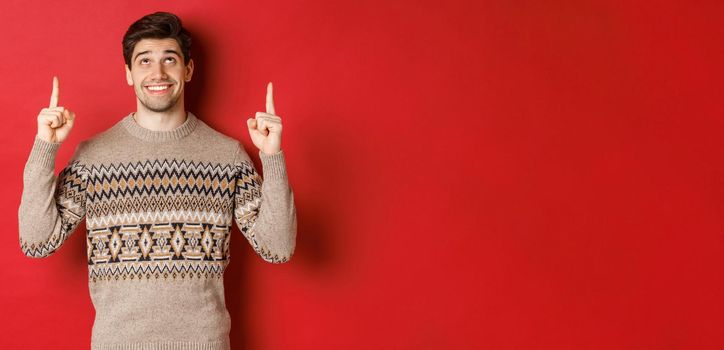 Portrait of dreamy handsome guy thinking about christmas holidays, wearing winter sweater, pointing and looking up at new year promo offer, standing over red background.