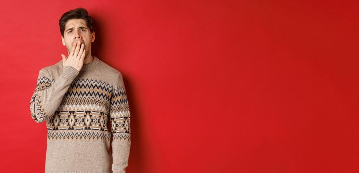 Image of tired or bored handsome man in winter sweater, yawning and covering mouth with hand, standing exhausted against red background.