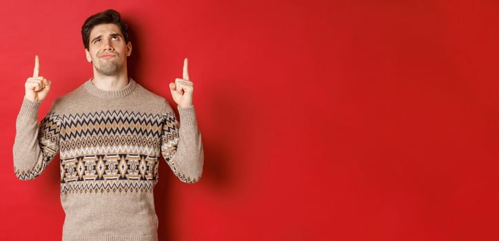 Image of disappointed and skeptical handsome man, wearing christmas sweater, smirk and frowning as looking at something bad, pointing fingers up, standing over red background.