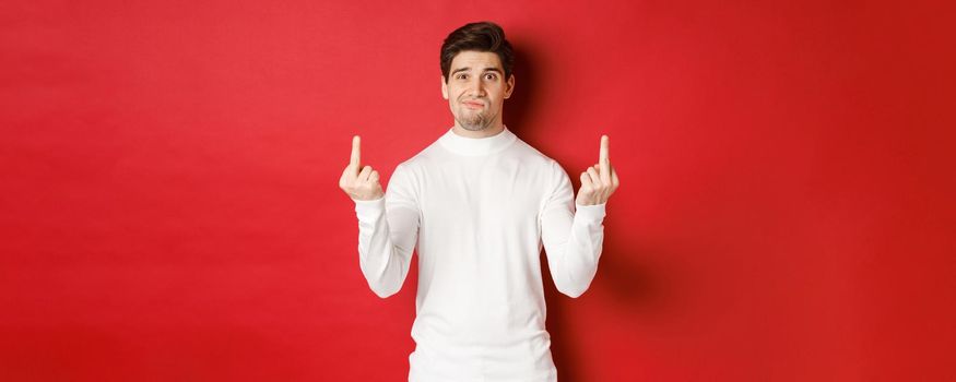Image of pissed-off and distressed man telling to fuck off, showing middle-fingers and looking upset, standing over red background in white sweater.