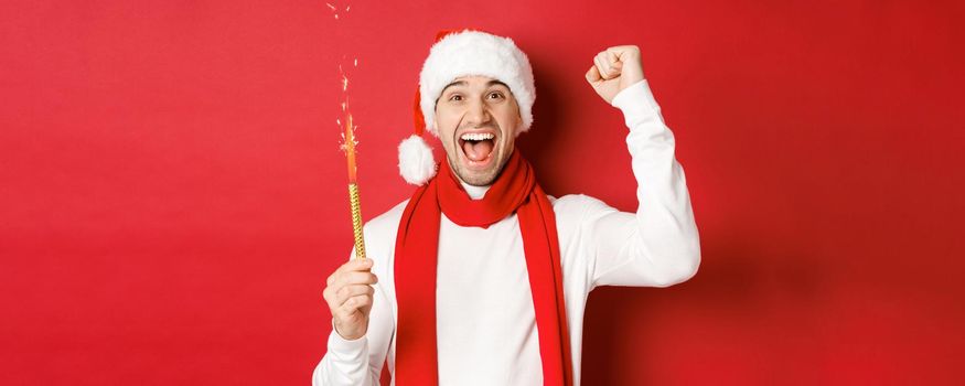 Concept of christmas, winter holidays and celebration. Handsome man celebrating new year and having fun, holding sparkler and smiling, wearing santa hat, standing over red background.