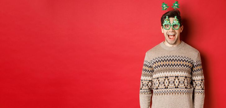 Portrait of surprised and excited handsome guy in party glasses and winter sweater, celebrating christmas and having fun, standing against red background.