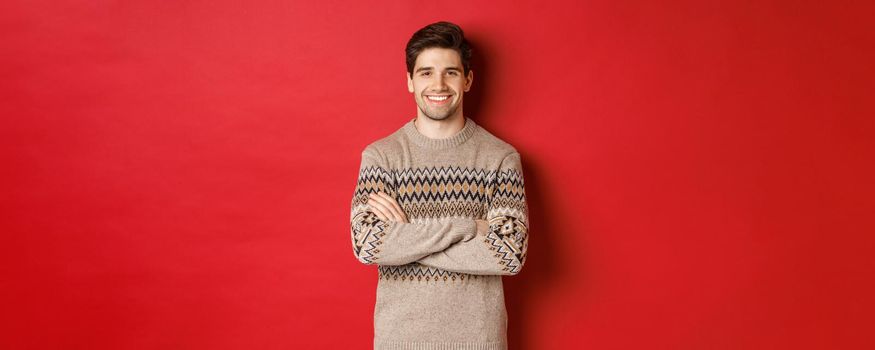 Image of handsome happy guy in christmas sweater, smiling and looking at camera, celebrating xmas holidays, standing over red background.