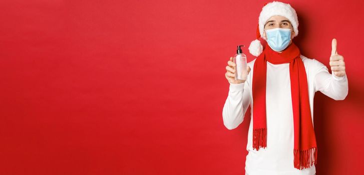 Concept of covid-19, christmas and holidays during pandemic. Happy and satisfied man in santa hat and medical mask, showing thumb-up in approval and recommending hand sanitizer.