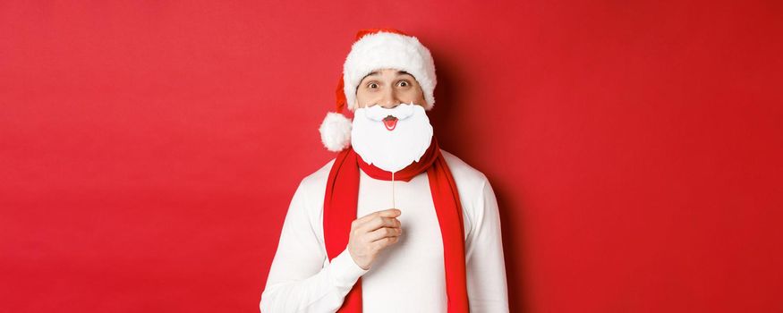 Concept of christmas, winter holidays and celebration. Portrait of funny man in santa hat, holding beard mask, enjoying new year party, standing over red background.