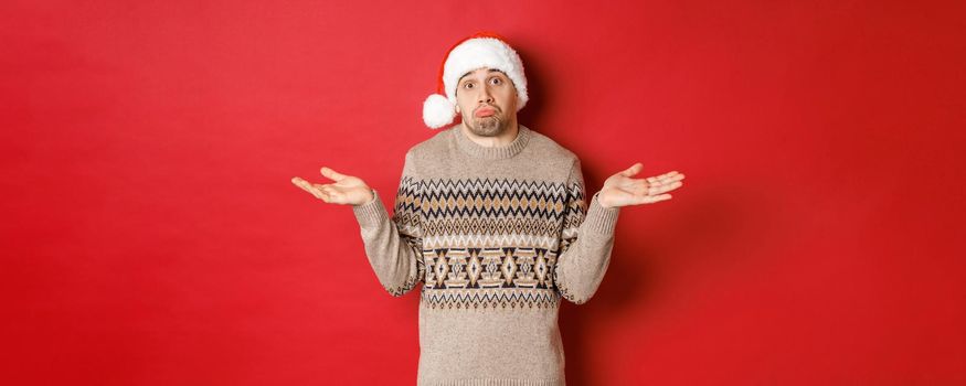 Image of clueless young man in sweater and santa hat, shrugging and looking unaware, dont know what to buy for new year holidays, standing over red background.