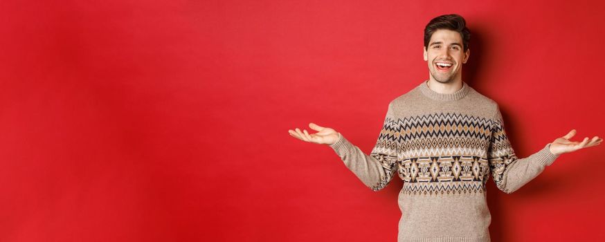 Portrait of happy good-looking man celebrating new year holidays, wearing christmas sweater, spread hands sideways and smiling, holding something on copy space, standing over red background.