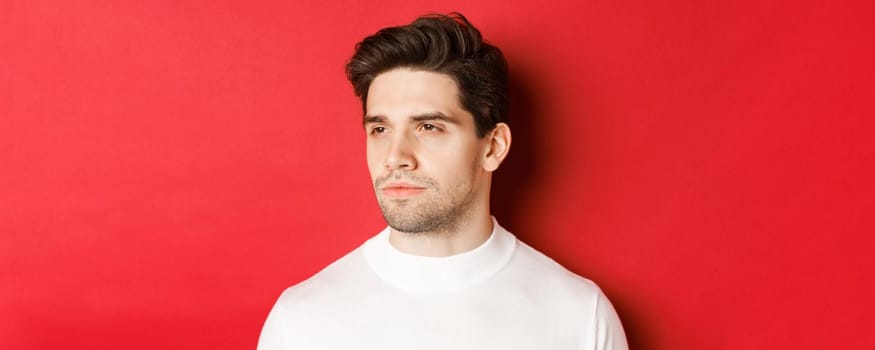 Close-up of serious and thoughtful man in white sweater, looking left, standing against red background.