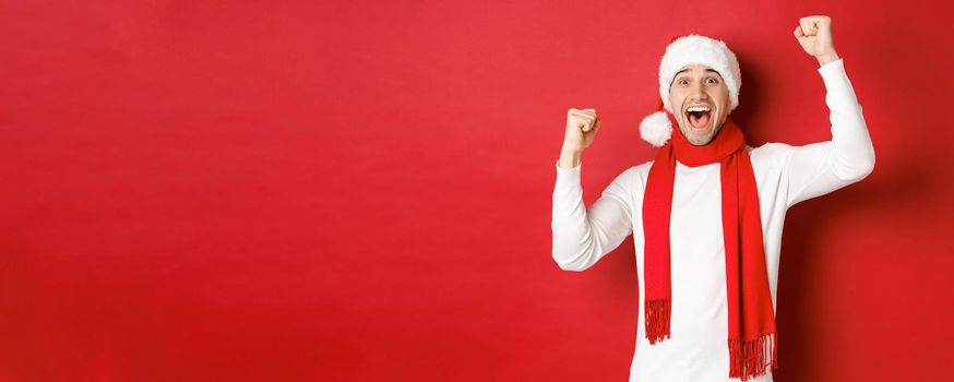 Image of joyful caucasian man in santa hat and scarf, shouting for joy and raising hands, celebrating victory or win, triumphing over red background.