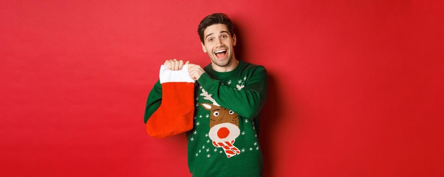Image of attractive cheerful man in sweater, holding christmas stocking with gifts, celebrating winter holidays, standing over red background.