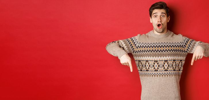 Concept of christmas celebration, winter holidays and lifestyle. Impressed handsome man in xmas sweater, pointing fingers down with amazed face, standing over red background.