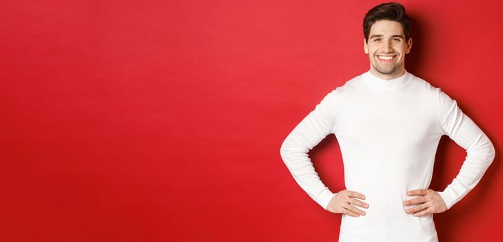 Portrait of handsome man with bristle, wearing white sweater, smiling and looking confident, standing against red background. Concept of new year and winter holidays