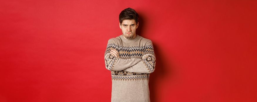 Image of angry and offedned guy in christmas sweater, feeling mad, cross arms on chest and sulking, standing over red background.
