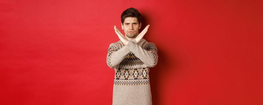 Image of angry and serious handsome man in christmas sweater, telling no or stop, showing cross gesture to restrict you from something, prohibit action, standing over red background.