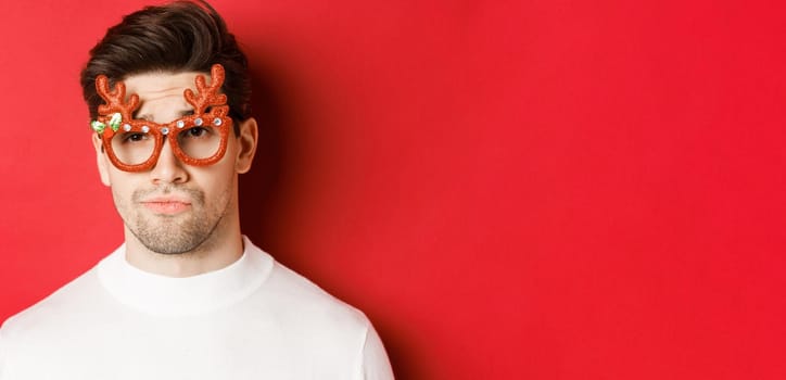 Concept of winter holidays, christmas and celebration. Close-up of skeptical handsome guy, wearing party glasses, smirk and looking unamused, standing over red background.