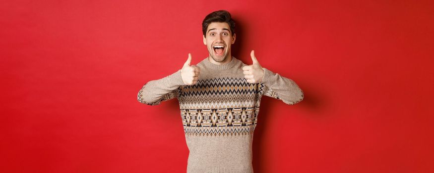 Portrait of cheerful man in christmas sweater like something good, showing thumbs-up and smiling amazed, praise new year party, standing over red background.