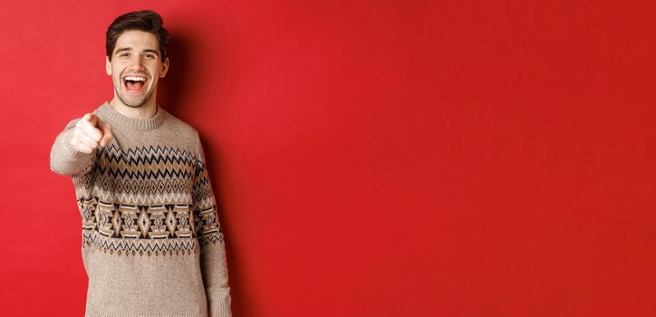 Concept of christmas celebration, winter and lifestyle. Happy attractive man in xmas sweater, wishing happy holidays and pointing at camera, standing over red background.