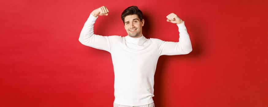 Portrait of smiling handsome man in white sweater, flexing biceps and bragging with strength, show-off strong muscles after workout, standing over red background.