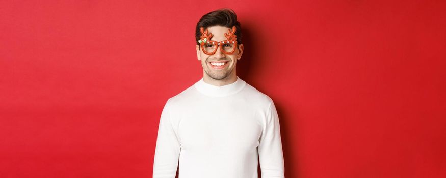 Close-up of attractive man in white sweater and party glasses, celebrating christmas, smiling amused, standing over red background.