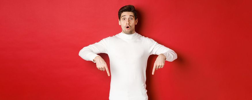 Image of surprised attractive guy in white sweater, showing announcement, pointing fingers down and looking amazed, standing over red background.