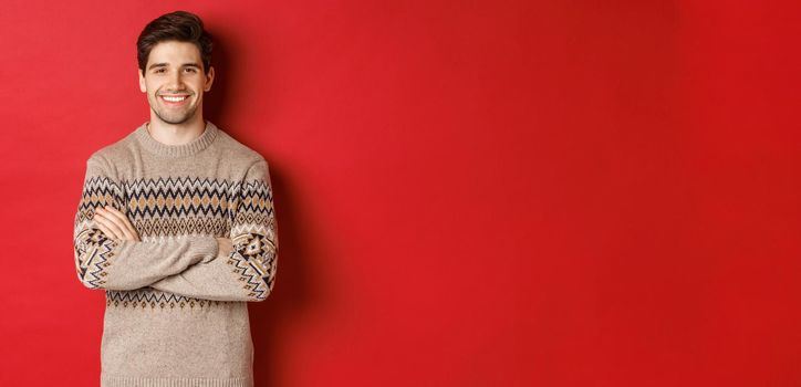 Image of handsome happy guy in christmas sweater, smiling and looking at camera, celebrating xmas holidays, standing over red background.