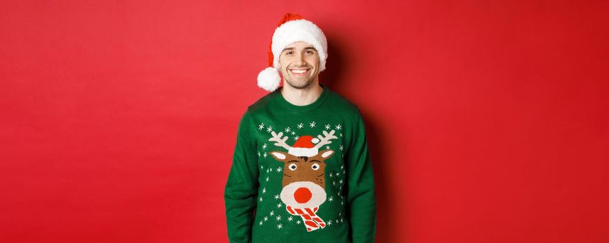 Concept of winter holidays, christmas and lifestyle. Handsome guy with bristle, wearing santa hat and green sweater, smiling joyful, celebrating new year, standing over red background.