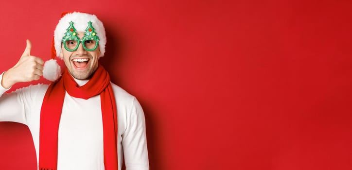 Concept of christmas, winter holidays and celebration. Cheerful guy in santa hat and party glasses, showing thumb-up and smiling pleased, standing over red background.