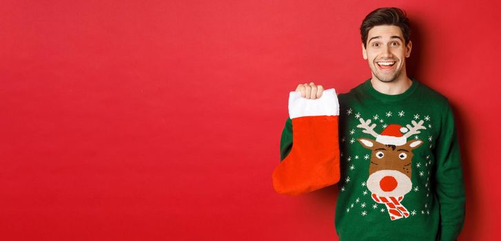 Image of handsome happy guy in sweater, holding christmas stocking and smiling amused, standing against red background.