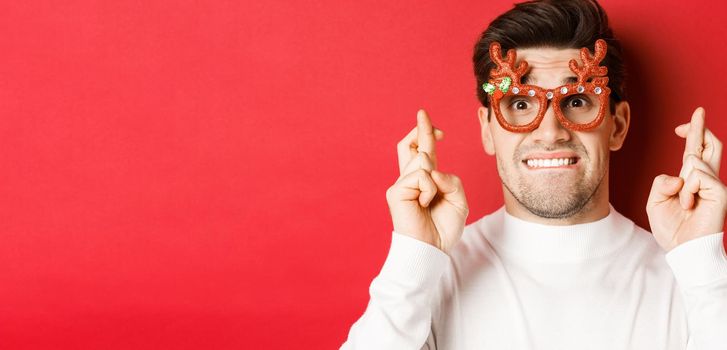 Concept of winter holidays, christmas and celebration. Close-up of nervous man in party glasses, crossing fingers for good luck and pleading, making a wish, standing over red background.