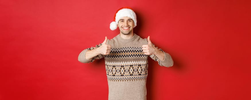 Image of cheerful attractive man in winter sweater and santa hat, showing thumbs-up, celebrating christmas and looking happy, like something, standing over red background.