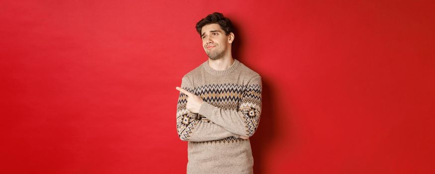 Image of disappointed handsome man in christmas sweater, looking and pointing left with upset face, express regret, showing new year advertisement, standing over red background.