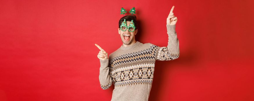 Portrait of joyful handsome guy in party glasses and christmas sweater, dancing and pointing fingers sideways, enjoying new year celebration, standing over red background.