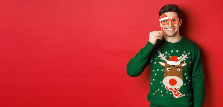 Portrait of handsome smiling man in christmas sweater and party glasses, celebrating new year and having fun, standing against red background.