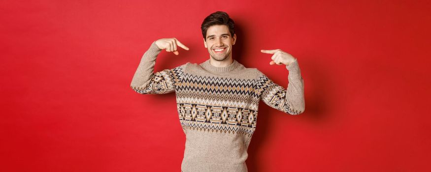 Concept of christmas celebration, winter holidays and lifestyle. Image of happy handsome man in xmas sweater pointing at himself and smiling, being secret santa, standing over red background.