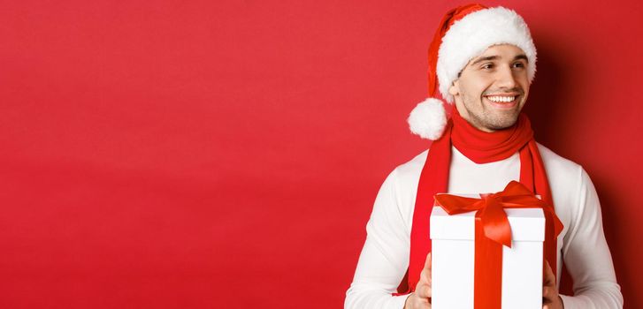 Concept of winter holidays, christmas and lifestyle. Close-up of attractive man in santa hat and scarf, holding new year gift, looking right and smiling, standing over red background.