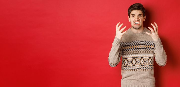 Image of angry and frustrated man in winter sweater, shouting and looking with hatred, standing upset over red background.